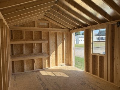 IMG_3866 - LELAND'S SHEDS - Interior of a partially built wooden shed with exposed studs, built-in shelves on the left, and a doorway opening to a sunny yard outside