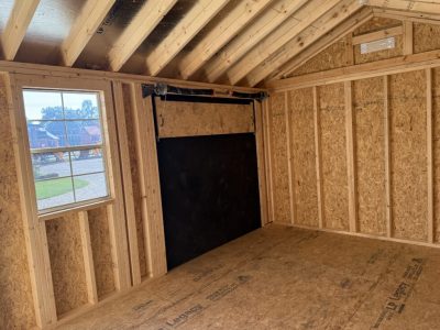 IMG_3868 - LELAND'S SHEDS - Interior view of a partially framed room with OSB walls, exposed wooden studs, a window, and a black sliding door panel/track.