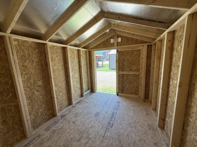 IMG_3873 - LELAND'S SHEDS - Interior of a small wooden shed under construction; exposed framing, OSB walls, foil-insulated roof, and an open doorway to outside.