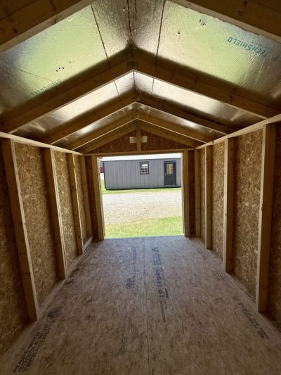 IMG_3874 - LELAND'S SHEDS - Interior of a small wooden shed under construction, with framed walls, foil insulation on the roof, and an open doorway showing a gray shed outside on a grassy lot.