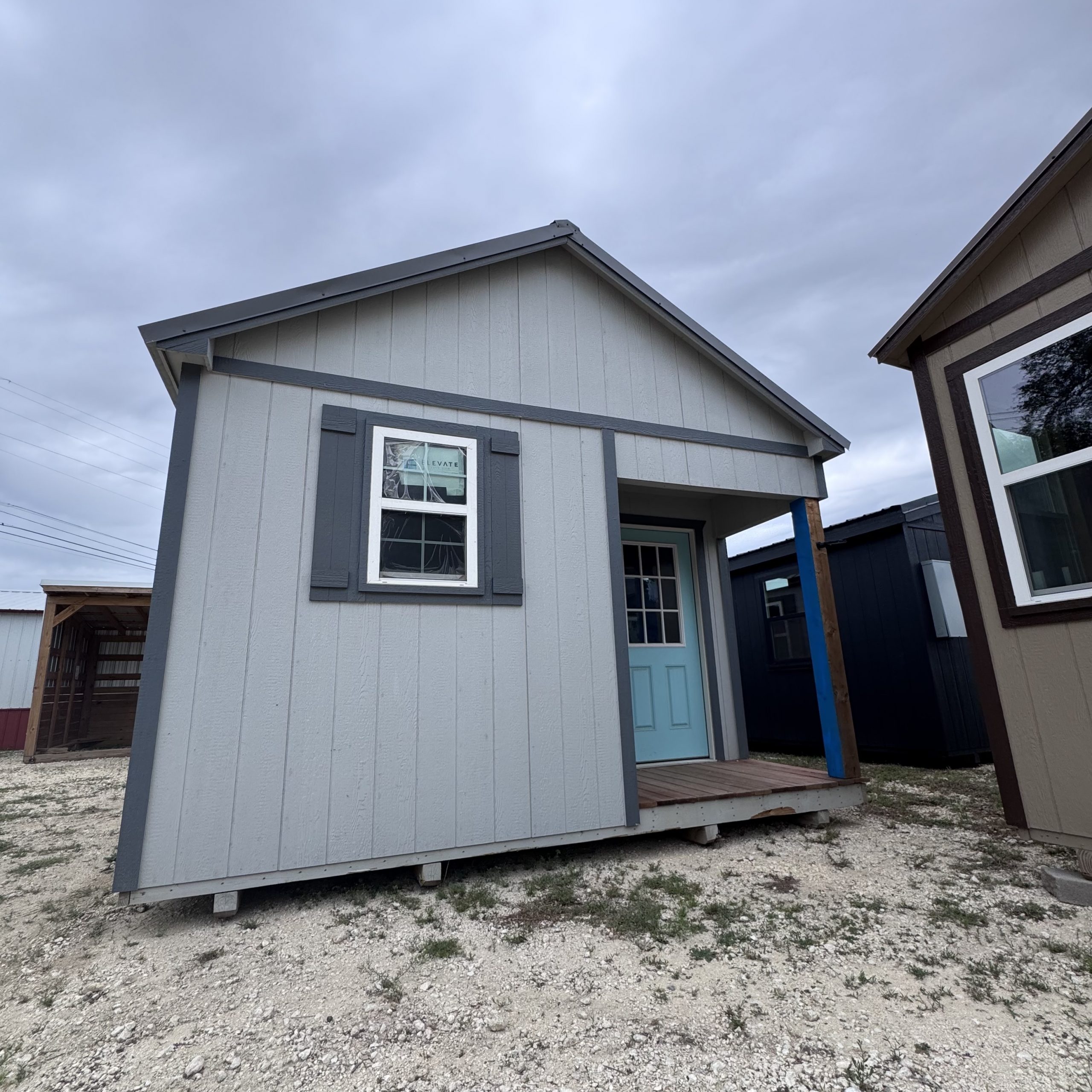 IMG_4063 - LELAND'S SHEDS - Corner view of a modern gray house with a gabled roof, window, and white trim; gravel yard in foreground under a cloudy sky.