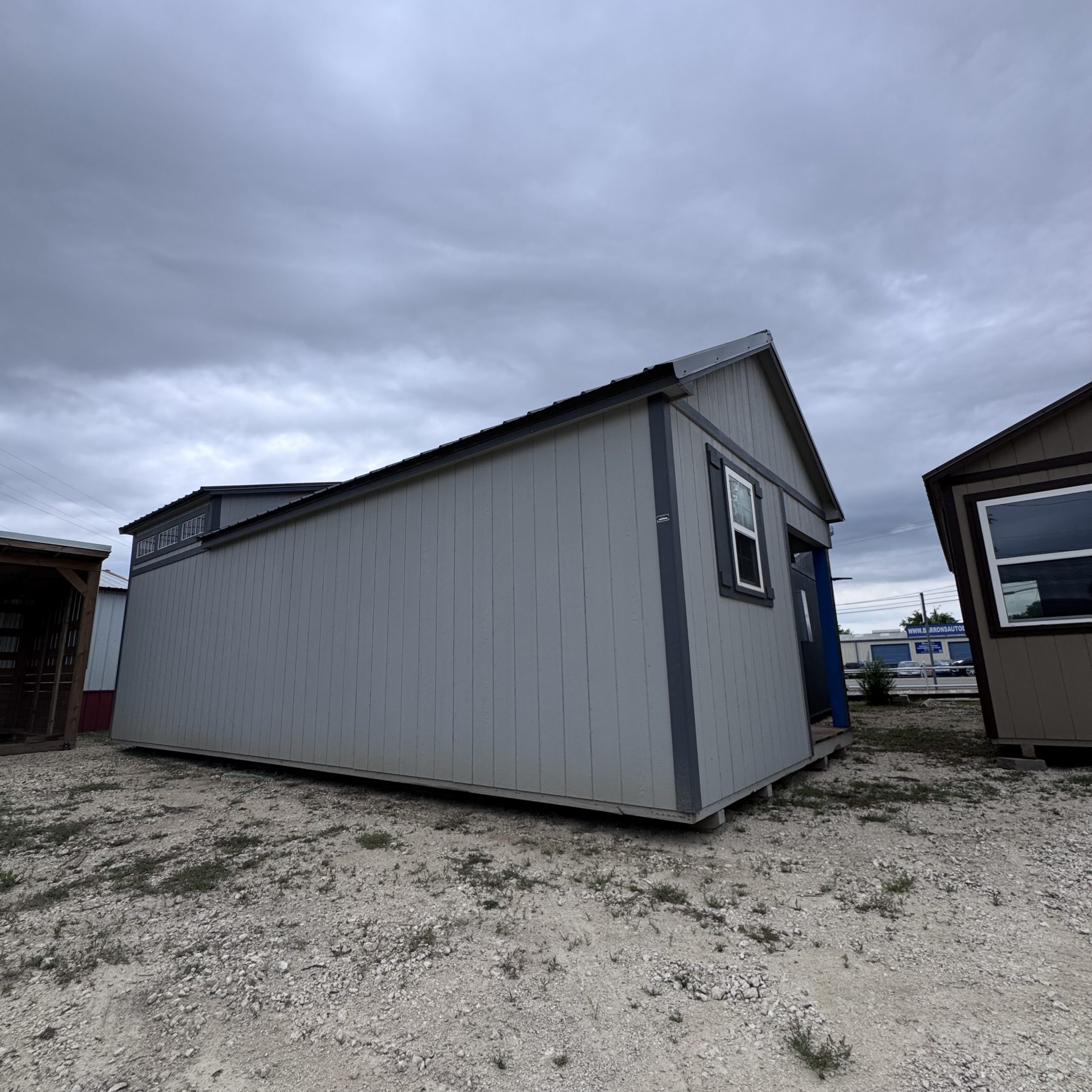IMG_4064 - LELAND'S SHEDS - Grey prefab modular building on a gravel lot under a cloudy sky.