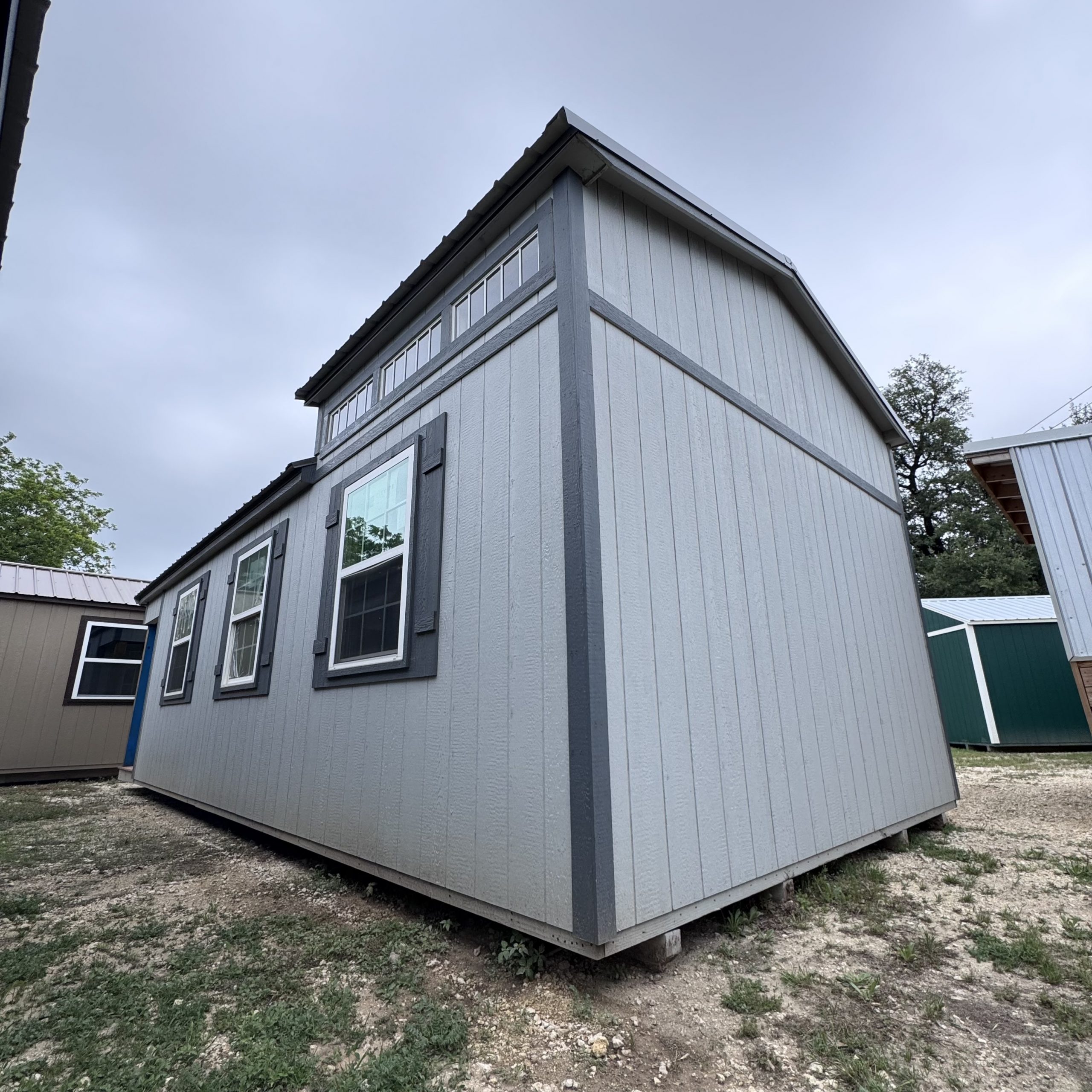 IMG_4065 - LELAND'S SHEDS - Corner view of a gray wooden house with vertical siding, dark trim, and a row of windows on a dirt/gravel lot.