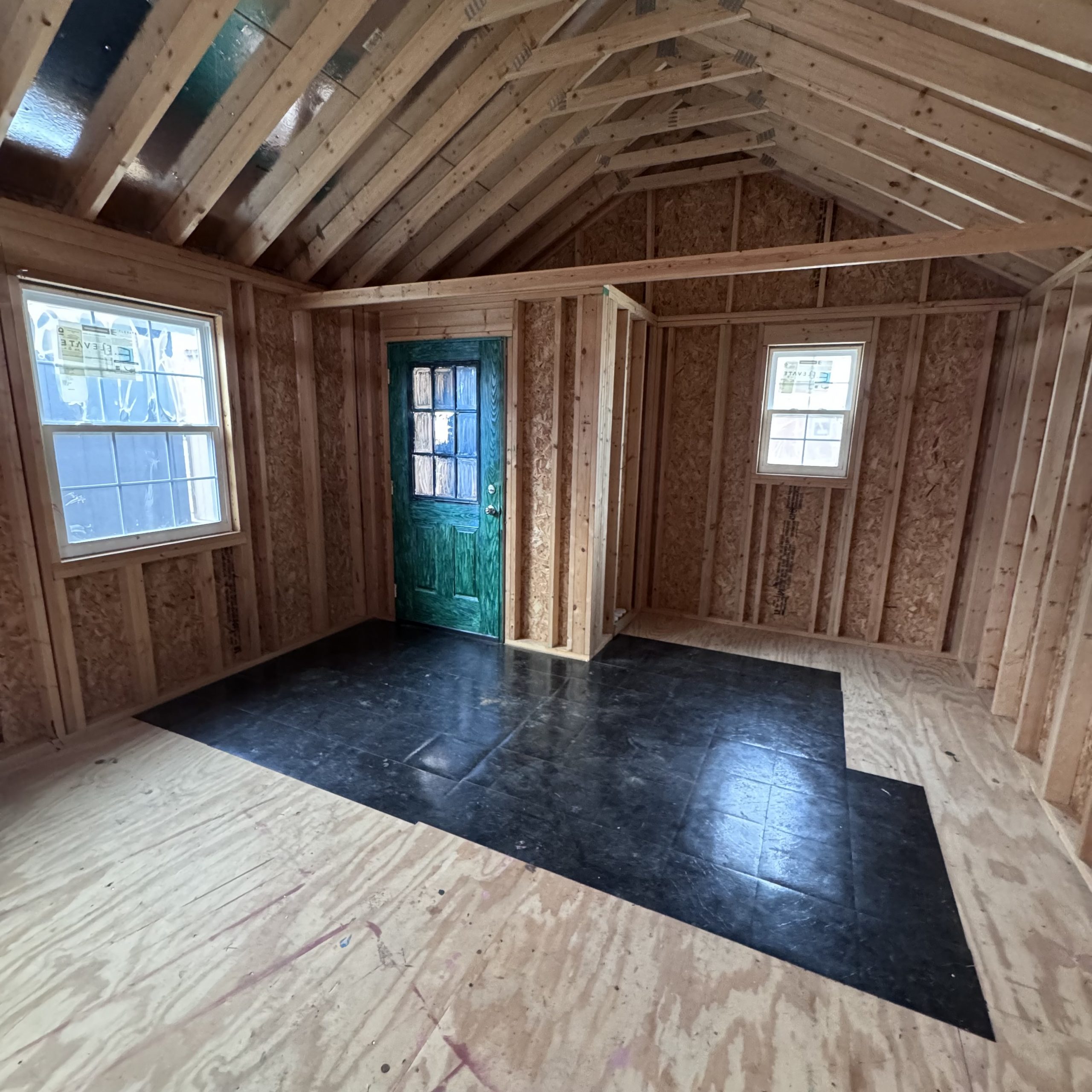 IMG_4070 - LELAND'S SHEDS - Interior of an unfinished room with exposed wooden framing, a green entry door, and several windows.