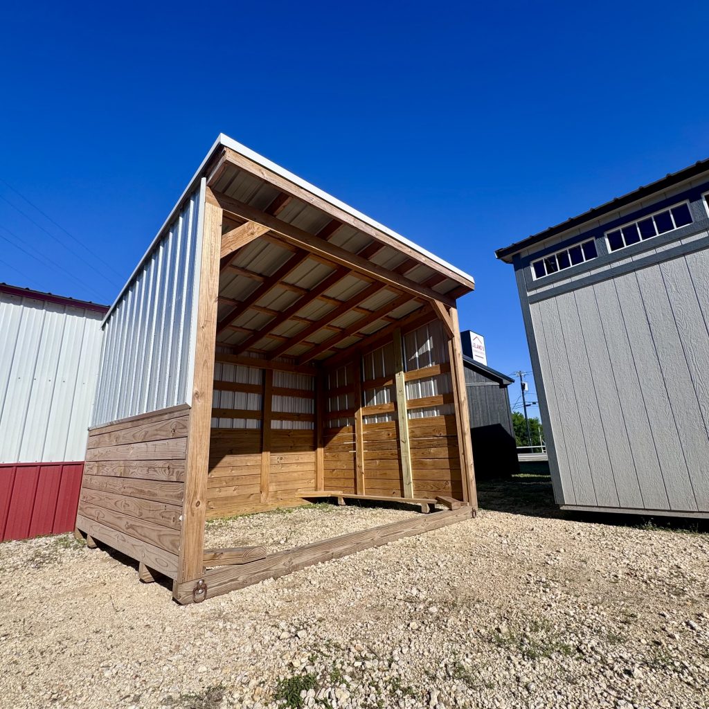 - - Wood-framed open shelter under construction beside a gray metal building against a clear blue sky