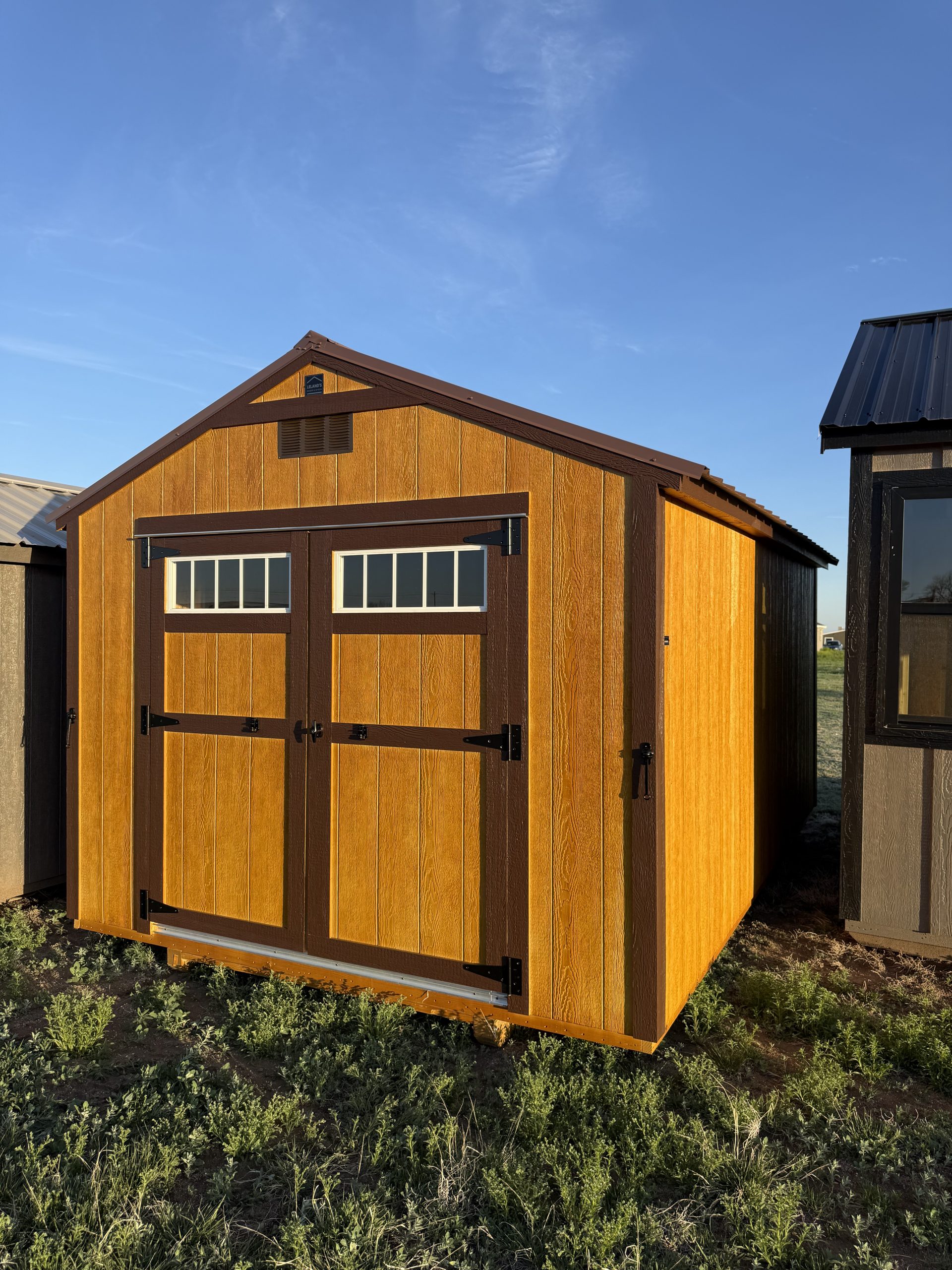  - Exterior view of a wooden shed with brown trim and a small windowed door on a grassy lot against a blue sky days.