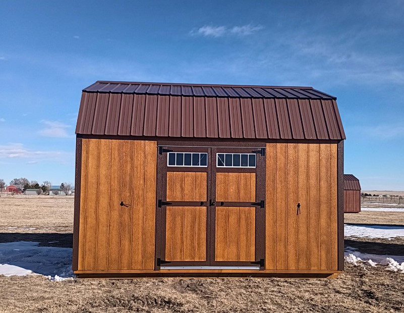  - Wooden shed with a brown metal roof in a flat field under a blue sky, featuring double doors with small window panels at the top.