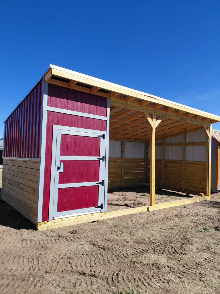  - Red barn-style shed with gray trim and a large door, beside an open wooden-framed shelter under a bright blue sky.