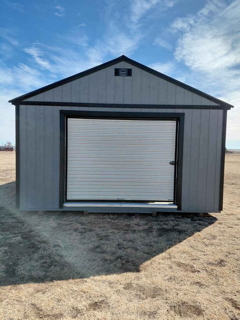  - Gray metal storage shed with a large white roll-up door in a dry grassy field under a blue sky, vent near the peak.