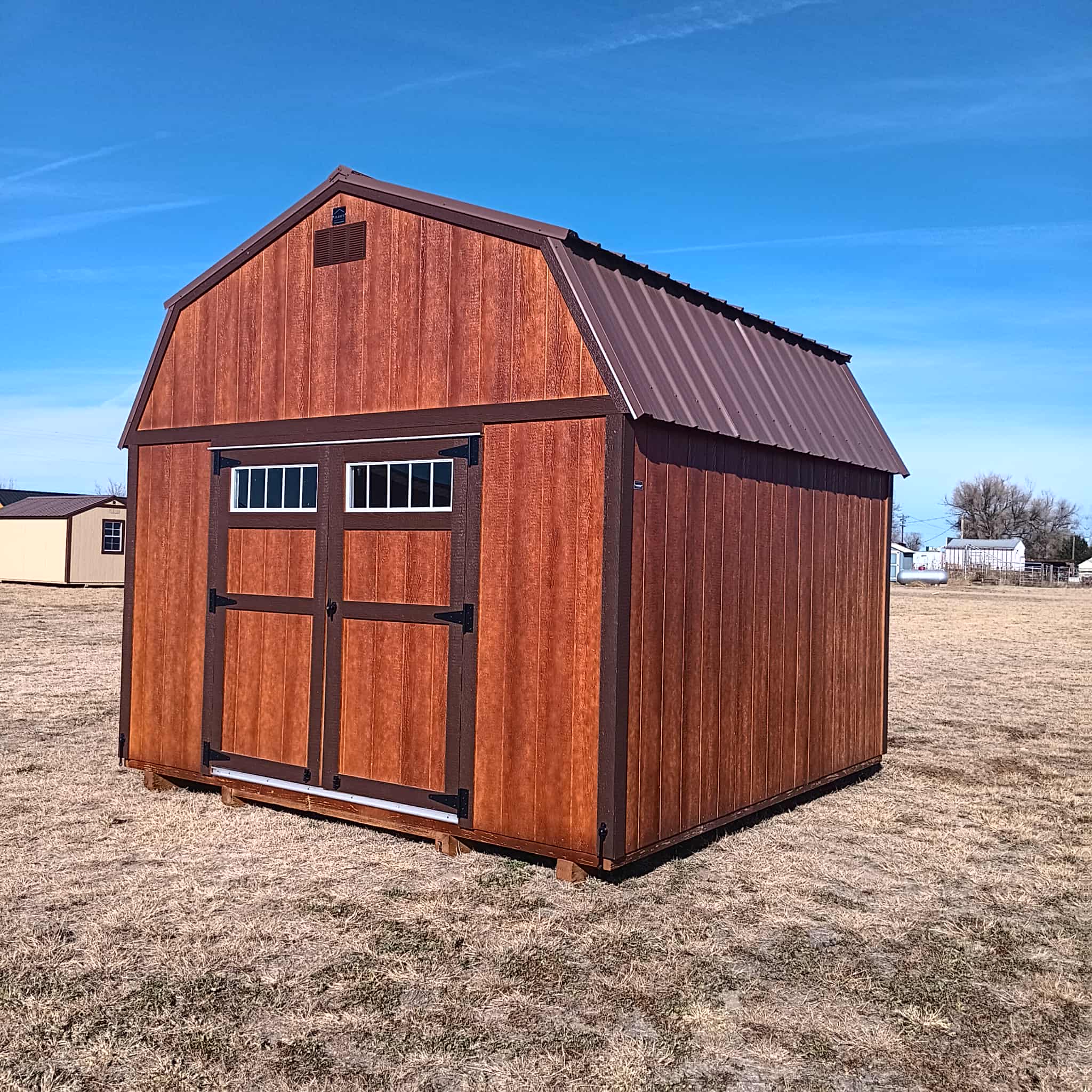  - Front view of a large red wooden barn with a dark metal roof and windowed wall, set in a dry, open field under a clear blue sky.