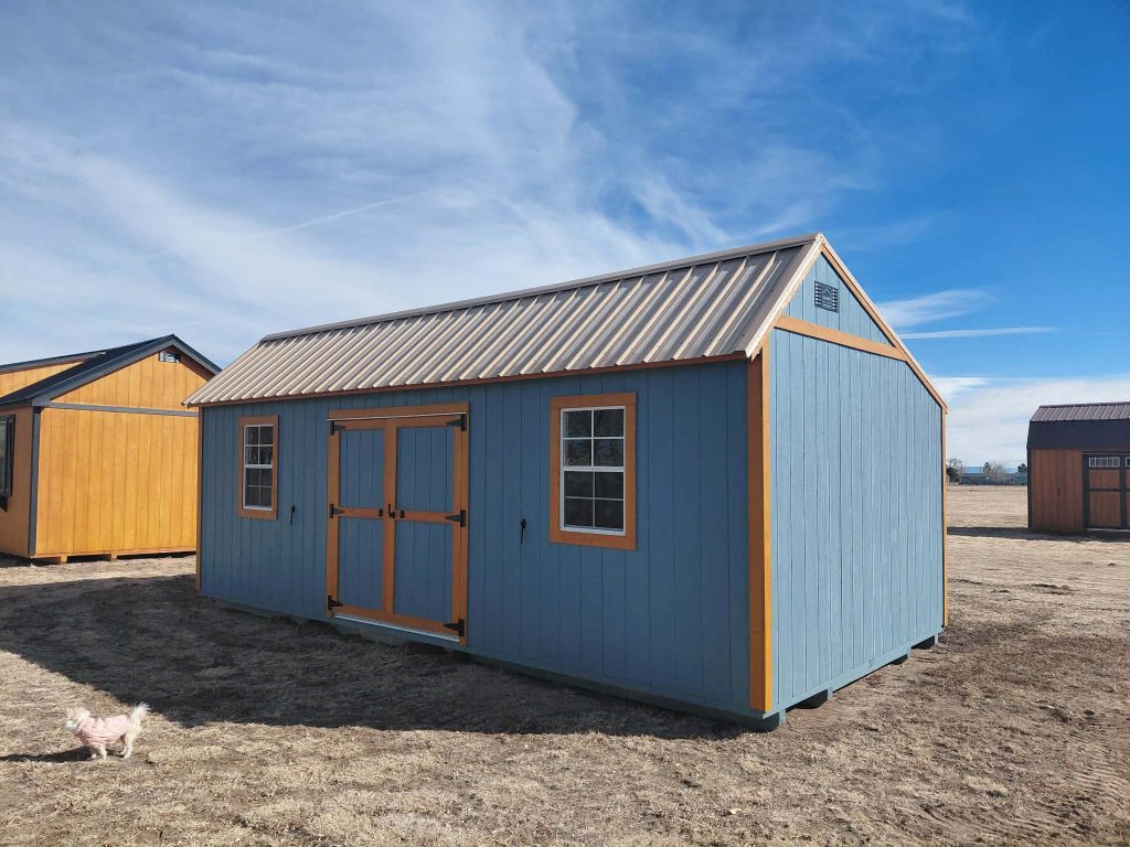  - Blue and orange wooden sheds sit on a dry field under a clear blue sky; a small white dog stands in the foreground left side.
