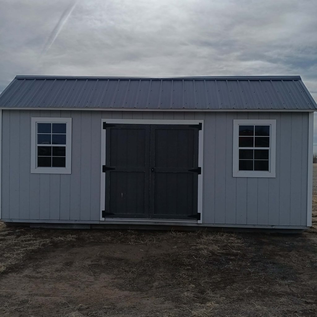  - Front view of a gray shed with a metal roof, two white-framed windows, and a dark central double door on dirt ground.
