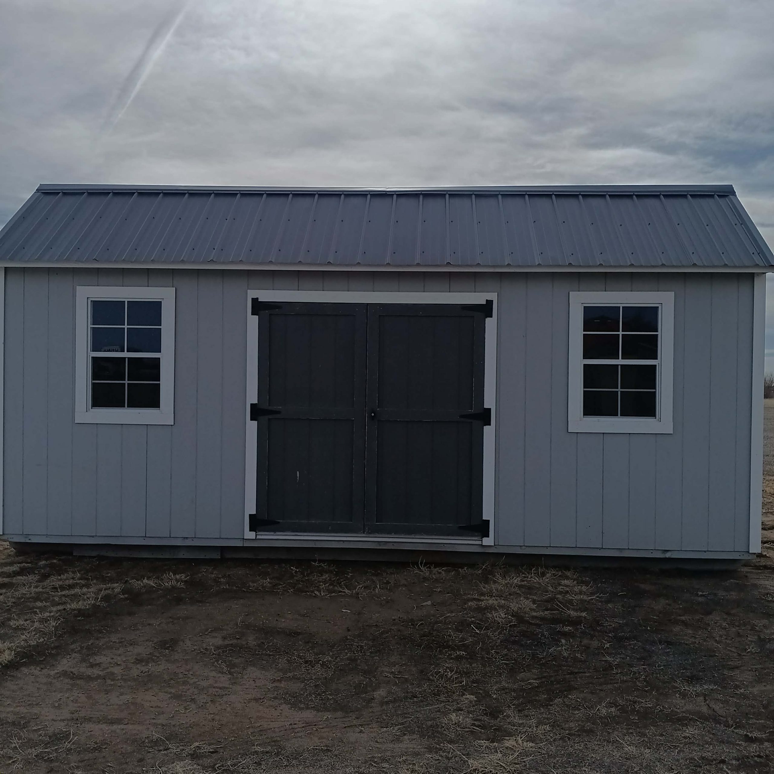  - Front view of a gray shed with a metal roof, two white-framed windows, and a dark central double door on dirt ground.