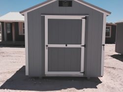 Sheds for Sale - Quality Guaranteed | LELAND'S SHEDS - Sheds for Sale - Quality Guaranteed | LELAND'S SHEDS - Small gray shed with white trim and a large double door in a dusty, sunny lot; other sheds visible in background.