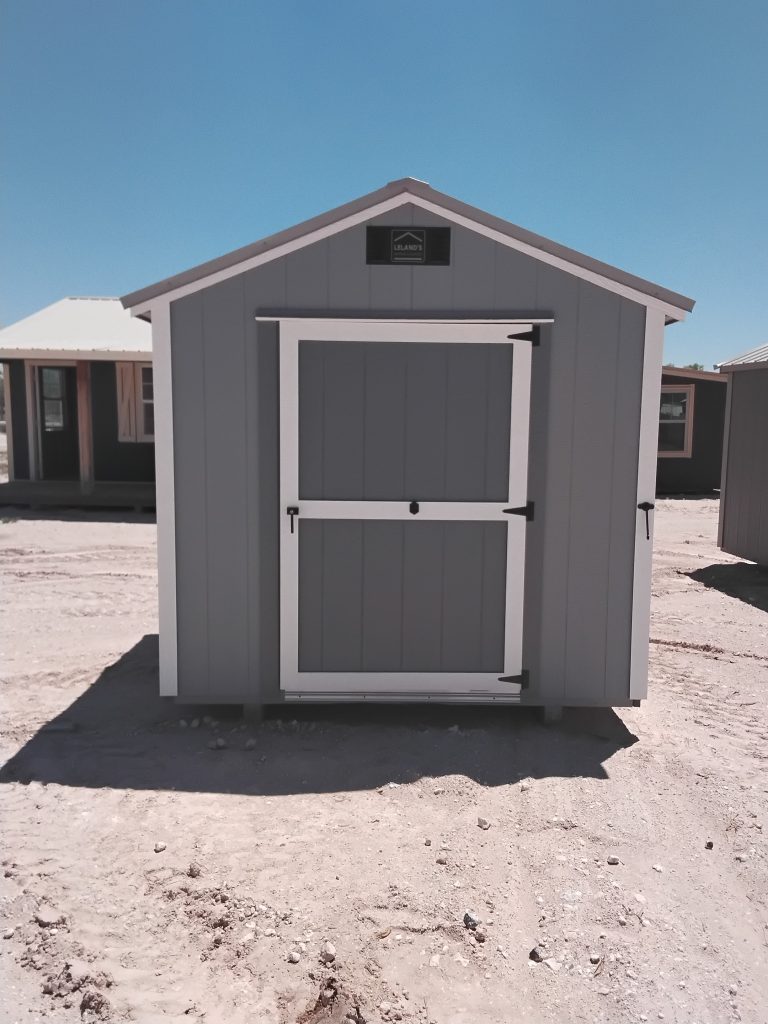 - - Small gray shed with white trim and a large double door in a dusty, sunny lot; other sheds visible in background.