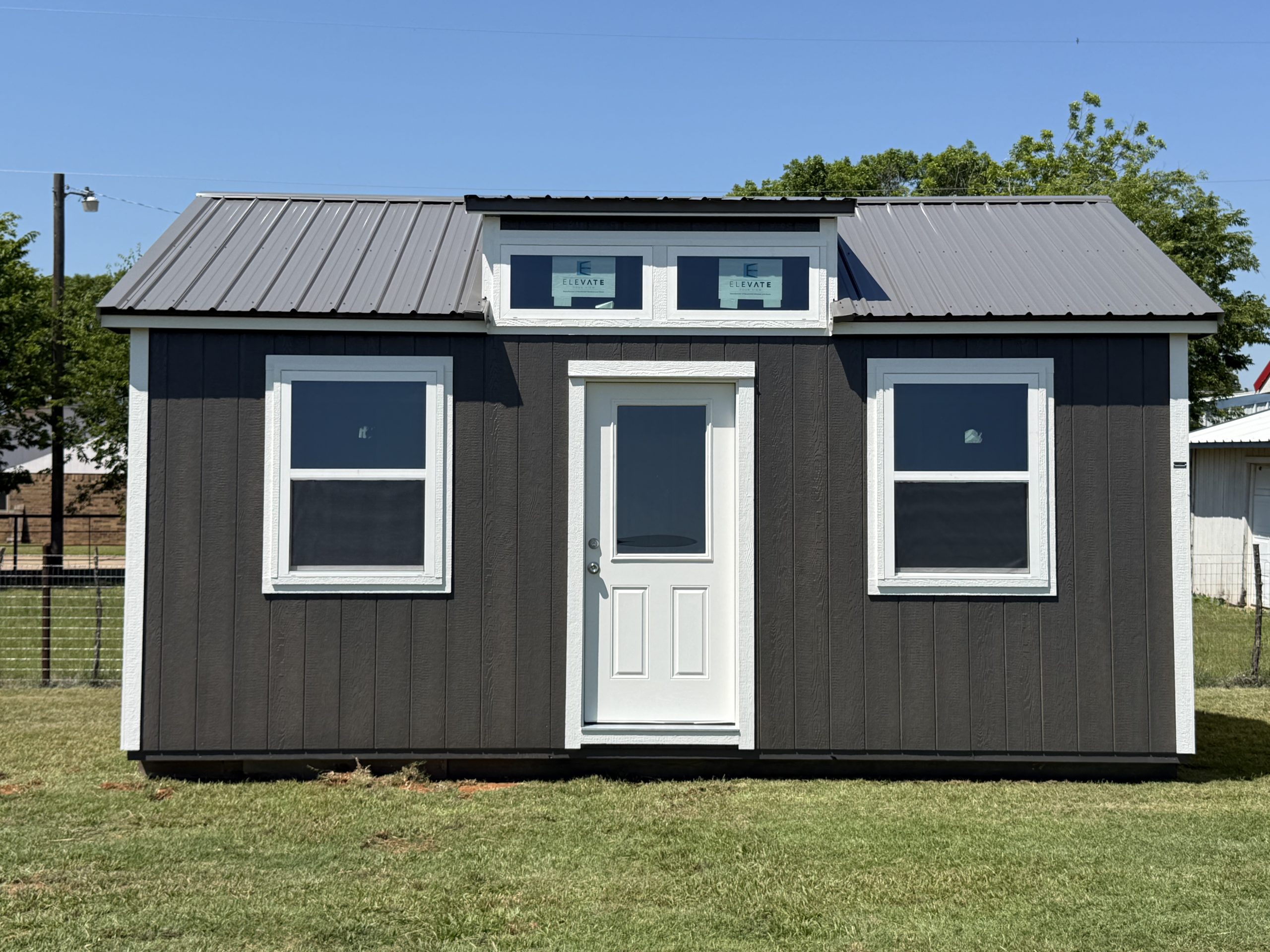 IMG_0224 - LELAND'S SHEDS - Front view of a small dark shed with white trim, metal roof, and two upper windows displaying 'Elevate' decals, on a grassy yard.