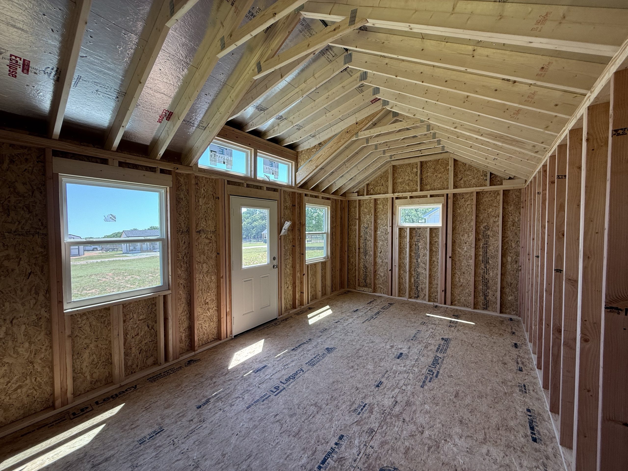 IMG_0229 - LELAND'S SHEDS - Interior of a house under construction: exposed wooden studs, roof trusses, and OSB sheathing with windows letting in daylight