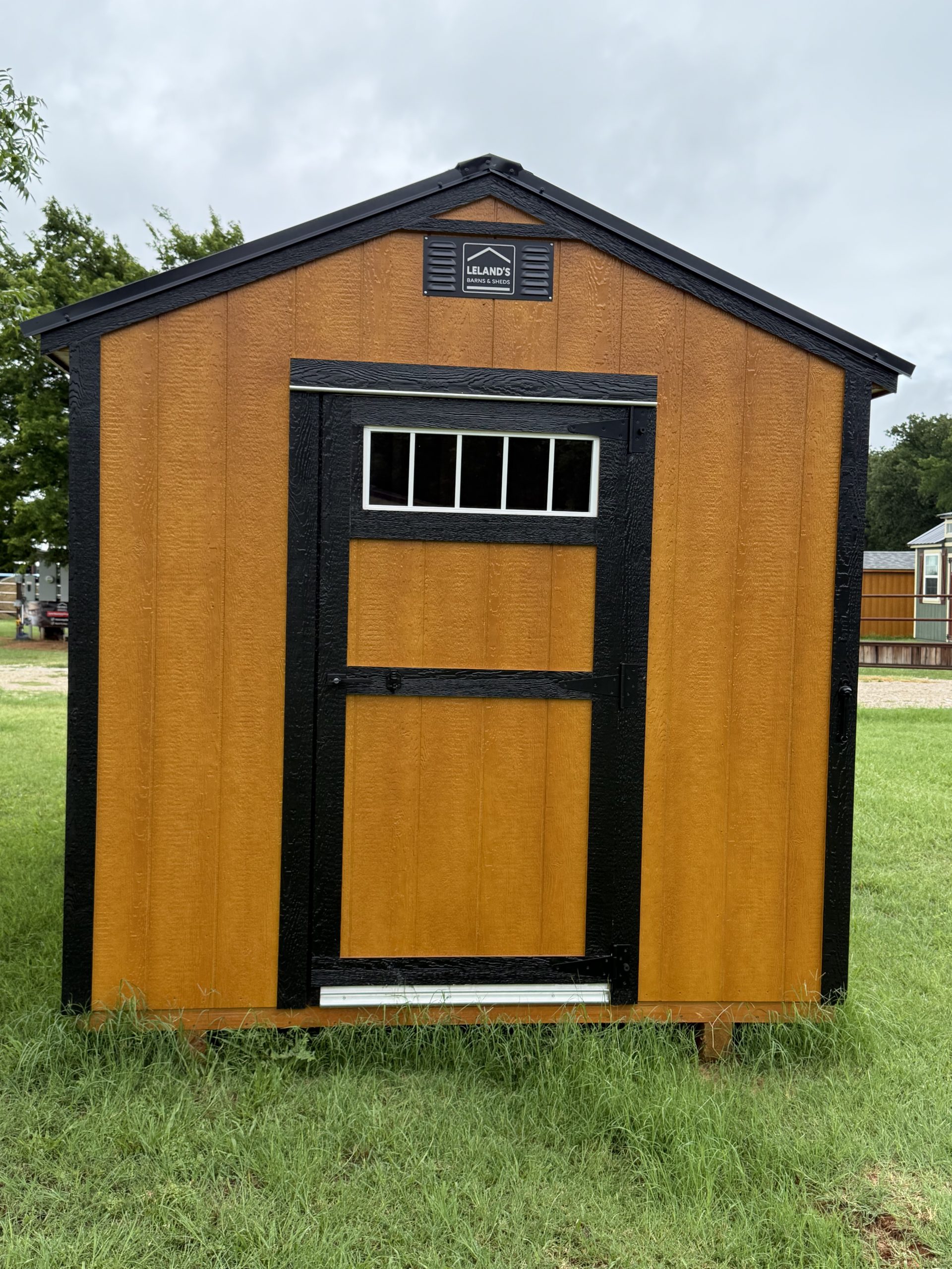 IMG_0850 - LELAND'S SHEDS - Front view of an orange shed with black trim, a small horizontal window, and a sign reading 'Leland's Barns & Sheds' at the top center.