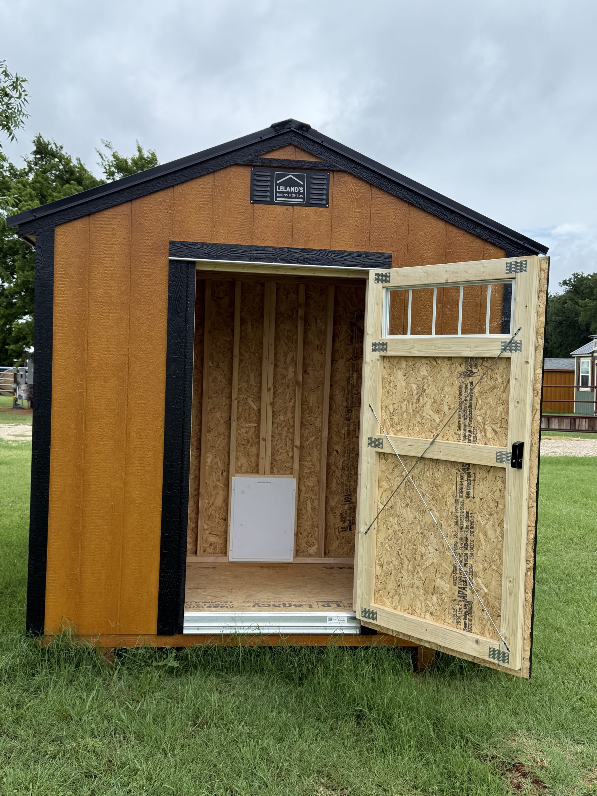 IMG_0851 - LELAND'S SHEDS - Small tan shed under construction with black trim, open front, and a detached door propped to the side; sign reads Leland's Barns & Sheds on the gable.