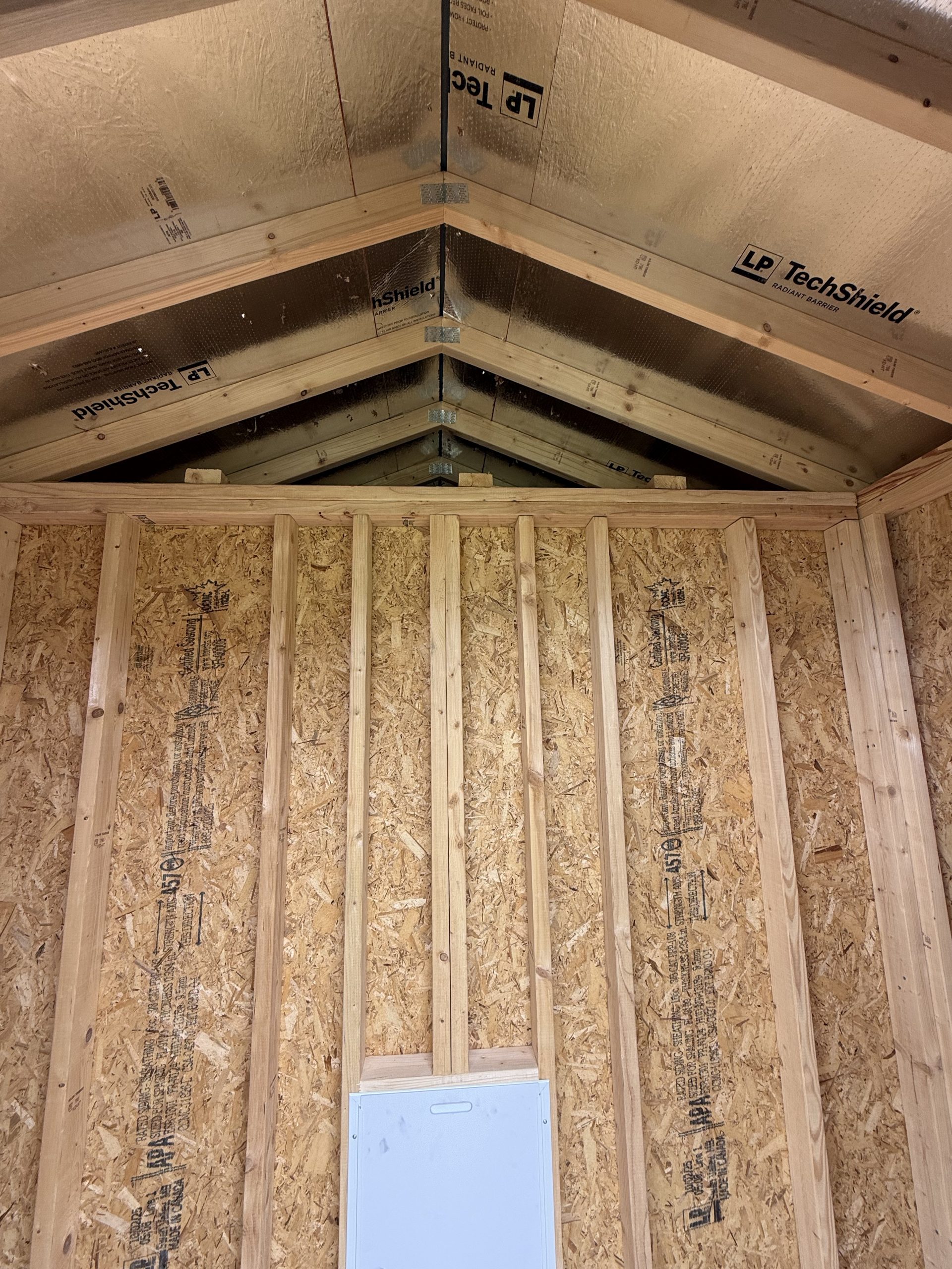 IMG_0852 - LELAND'S SHEDS - Attic framing with exposed wooden rafters and foil-backed insulation on the roof, OSB walls, and a white electrical panel near the bottom central area.