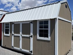 Sheds for Sale - Quality Guaranteed | LELAND'S SHEDS - Sheds for Sale - Quality Guaranteed | LELAND'S SHEDS - Brown shed with white-framed windows and a white metal roof on a gravel lot, clear blue sky.