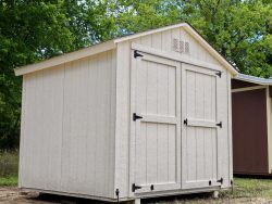 Sheds for Sale - Quality Guaranteed | LELAND'S SHEDS - Sheds for Sale - Quality Guaranteed | LELAND'S SHEDS - Beige wooden shed with a vent and black hinges, outdoors among green trees and blue sky.