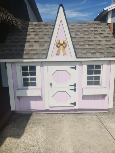 A pink and white 8x8 Victorian Playhouse with a bow on the roof, available at sheds on sale.