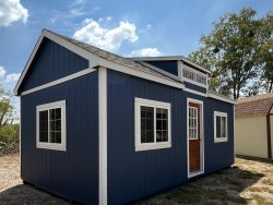 A blue and white 12x24 Chalet Shed, resembling a shed on sale, sitting on a dirt lot.