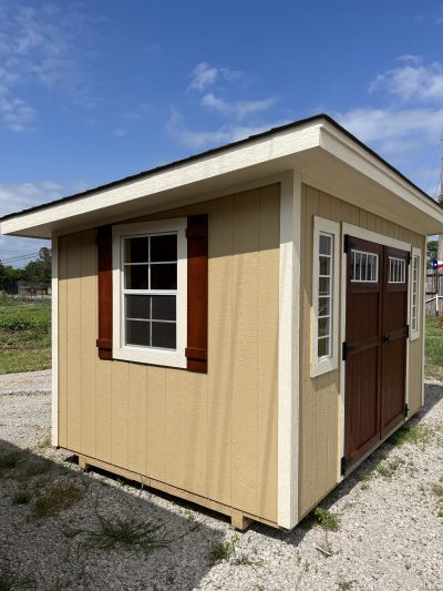 A small 8x12 Studio Shed with tan siding and brown shutters, available for sale at a nearby shed store.