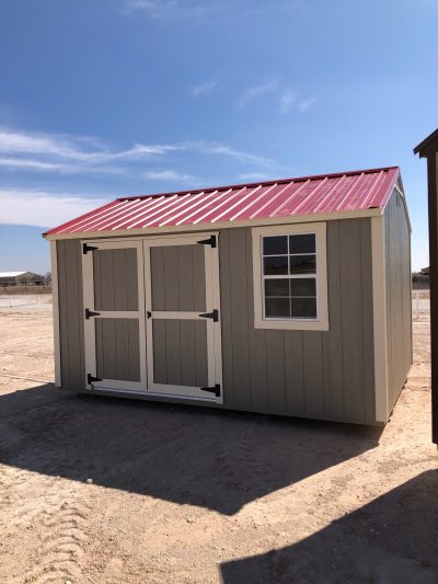 A red-roofed 10x14 Utility Shed in the middle of a dirt lot, available for sale.