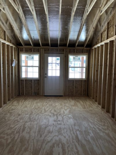 An empty room with wood framing, windows, and a 12x28 Cottage Shed on sale nearby.
