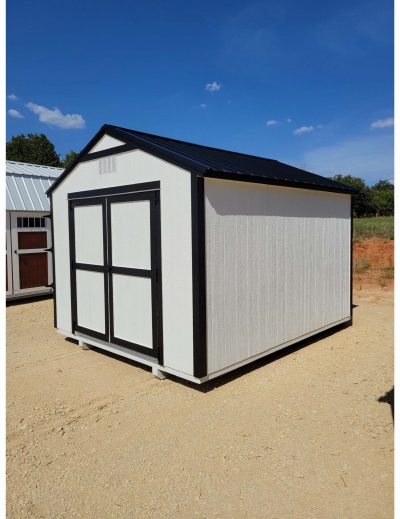 A white and black 10x12 Utility Shed for sale sitting on a dirt lot near a shed store.